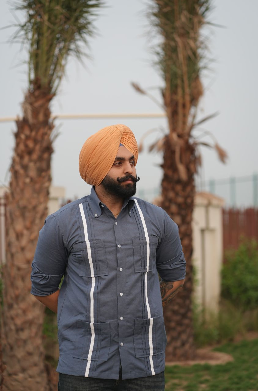 Man wearing a blue shirt with white stripes and an orange turban standing outdoors with palm trees in the background.