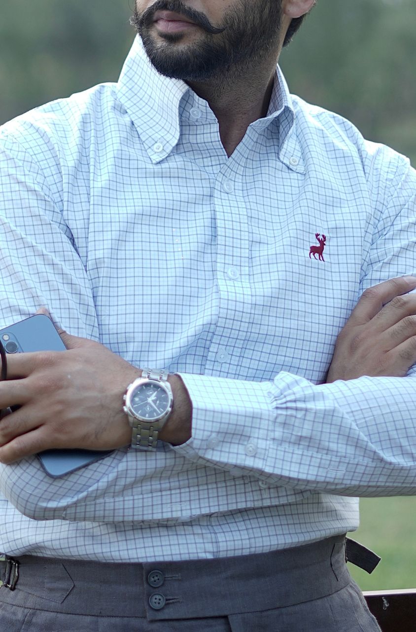 Man wearing a light blue checkered shirt with a small red logo, holding a phone, and wearing a watch.