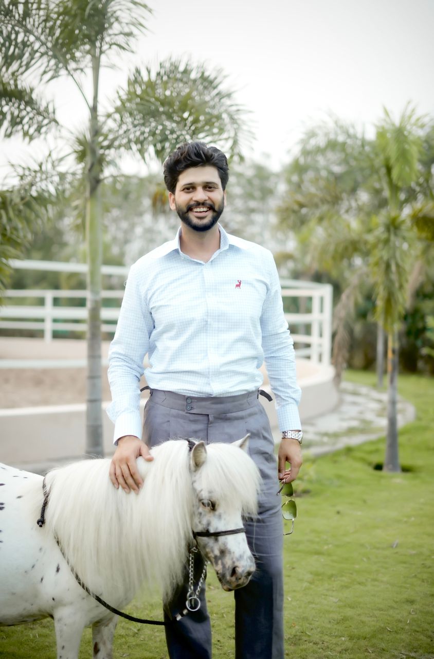 Man standing next to a small white horse in an outdoor setting with greenery.