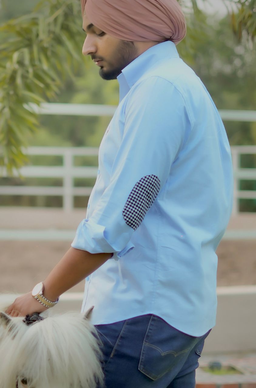 Man wearing a light blue shirt with rolled-up sleeves and a pink turban, standing outdoors.
