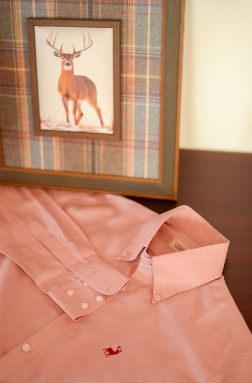 Pink shirt with a small logo on a table, framed picture of a deer on plaid wall.